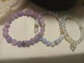 Trio of layered stretch bracelets in lavender, blue-white, and opalescent beads with silver accents and a heart-cross charm.