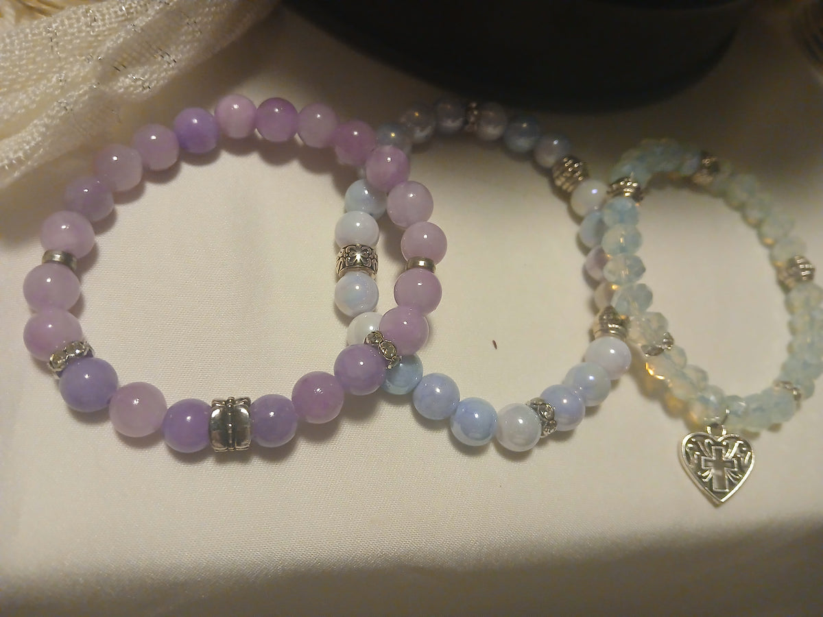 Trio of layered stretch bracelets in lavender, blue-white, and opalescent beads with silver accents and a heart-cross charm.