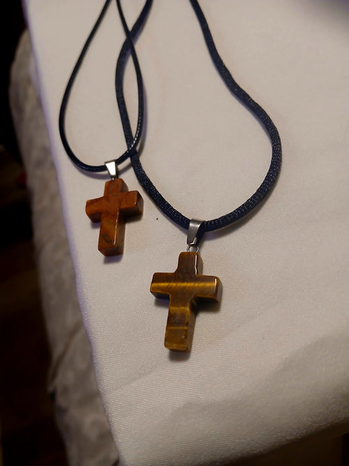 Two polished stone cross style necklaces with black cords. One features golden tiger eye, the other reddish brown stone, both with silver bail connectors.